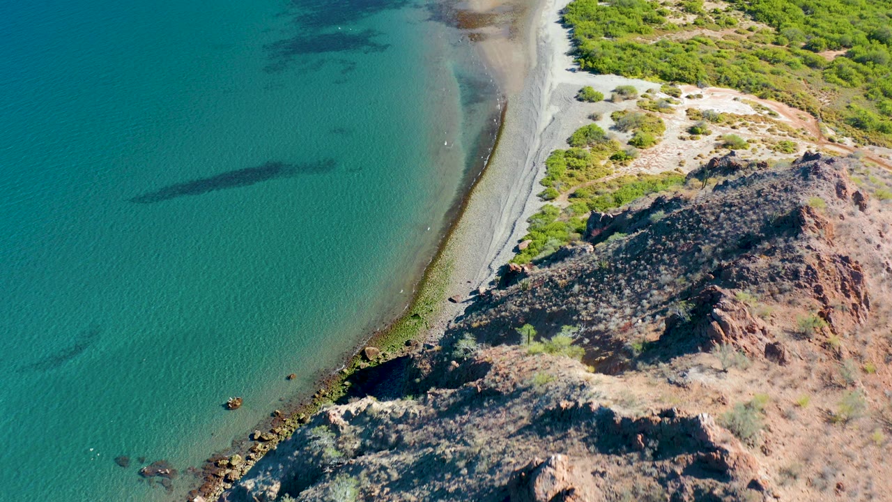 Aerial view in Ligui, Baja California Sur shows the beach running diagonally between turquoise water and a rocky scrub-covered slope, with no visible people.