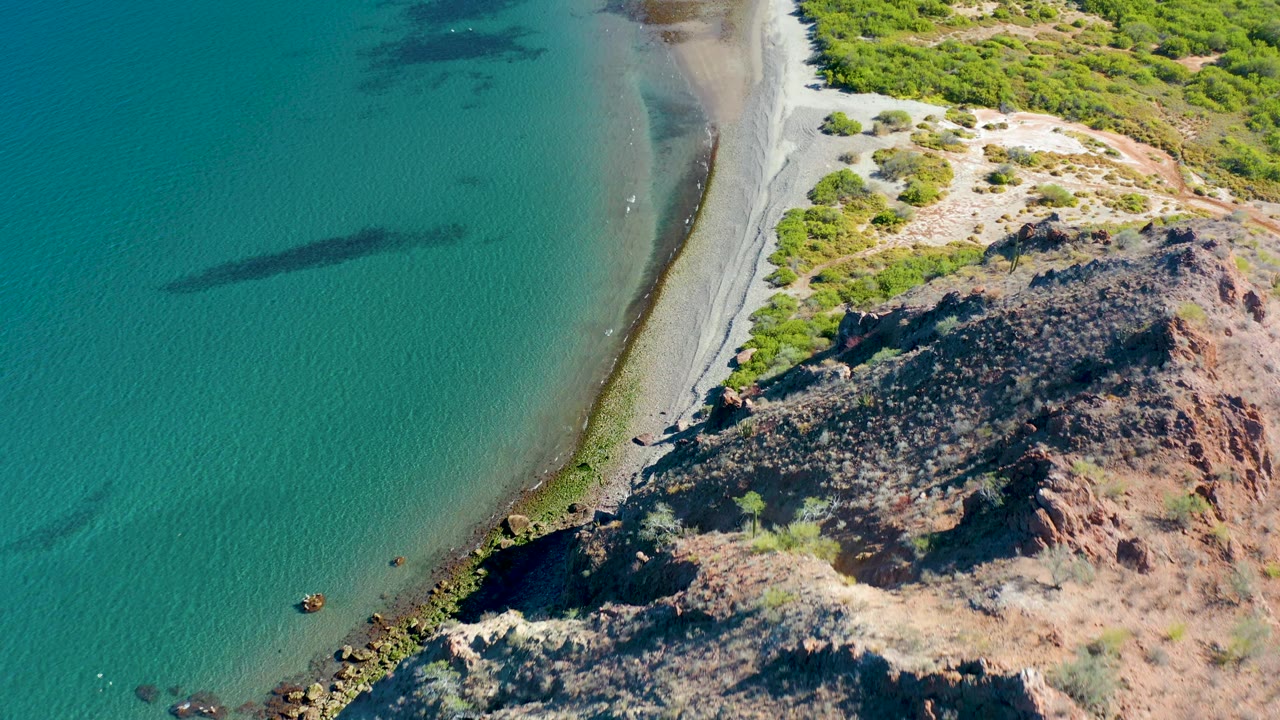 High oblique frame over Ligui, Baja California Sur shows blue-green water to the left, a narrow pale shoreline, and dark rocks with low vegetation to the right, with no visible people.