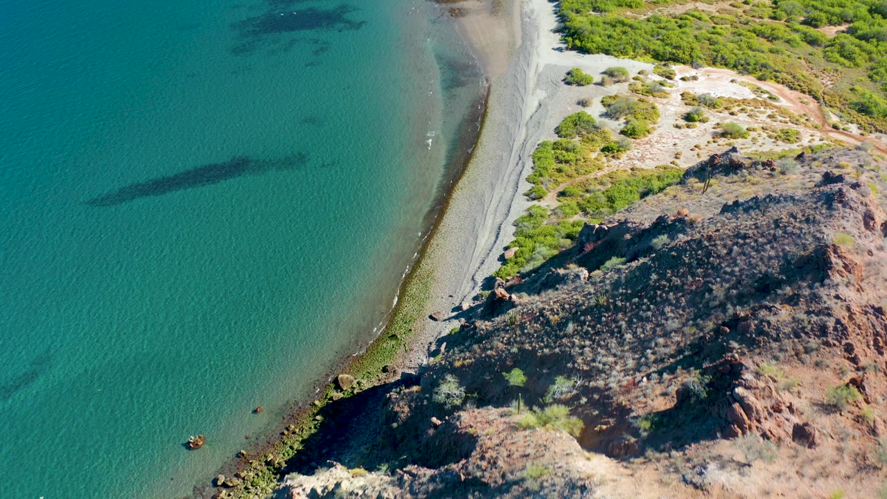 Aerial frame in Ligui, Baja California Sur shows calm turquoise water beside a thin beach line, with jagged rocks and scrub forming the land side, with no visible people.
