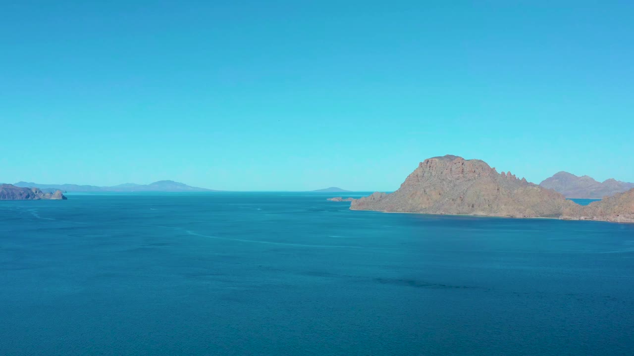 Wide aerial view over blue sea toward a rocky island near Ligui, Baja California Sur, with distant landforms on the horizon and no visible people.