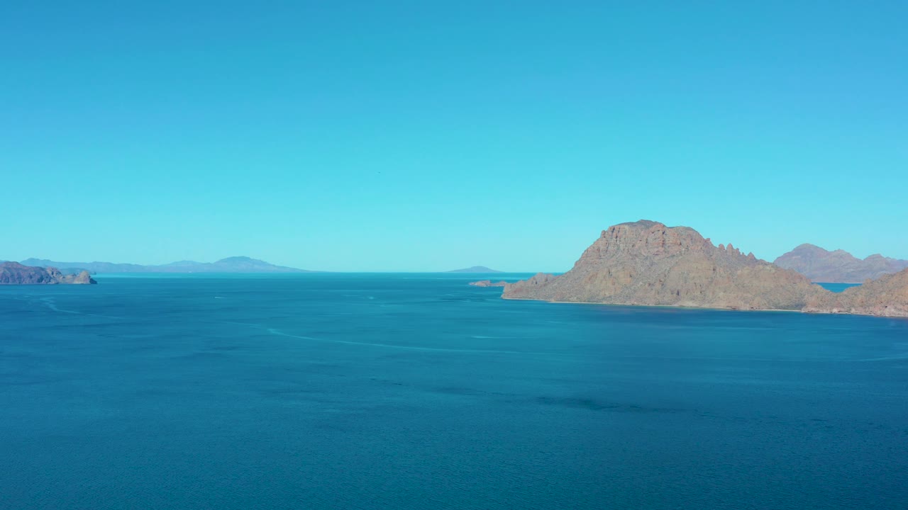A broad ocean surface leads to a low rocky island and distant shoreline shapes under clear cyan sky in Ligui, Baja California Sur, with no visible people.
