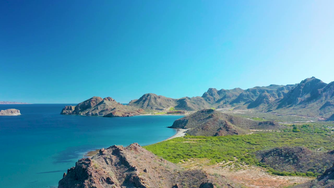 Opening frame of Ligui, Baja California Sur showing turquoise sea beside rocky cliffs, a pale sandy beach, green coastal vegetation, rugged mountains, clear sky, and no visible people.