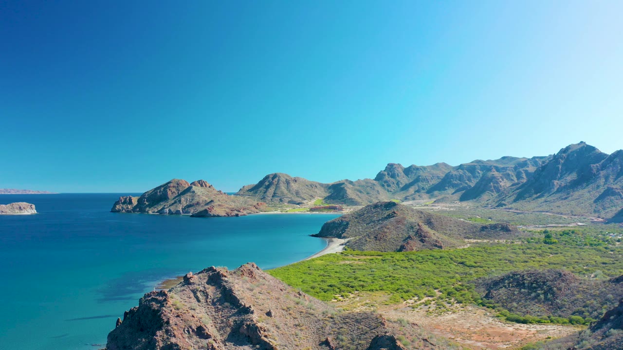 A bright Ligui bay view with turquoise water, a rocky foreground slope, sandy shoreline, green vegetation across the flat terrain, distant mountains, and no visible people.