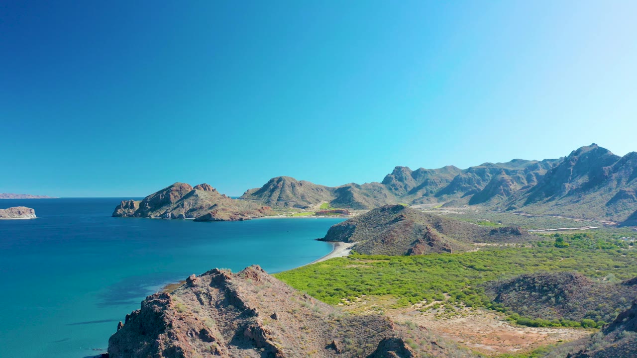 Coastal panorama with calm sea, rugged rocks, beach, vegetation, and mountains