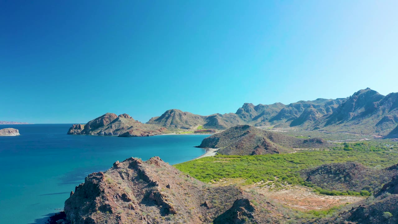 Aerial view in Ligui, Baja California Sur, with the foreground rock slightly larger, blue-green ocean along the left edge, a pale beach in the distance, mountains across the background, and no visible people.