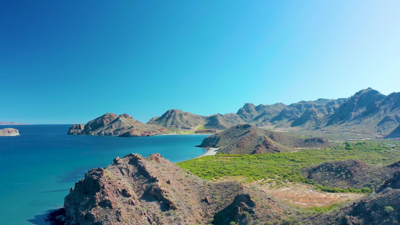 Wide aerial coastline with foreground rocks, calm sea, sandy cove, and dry mountains.