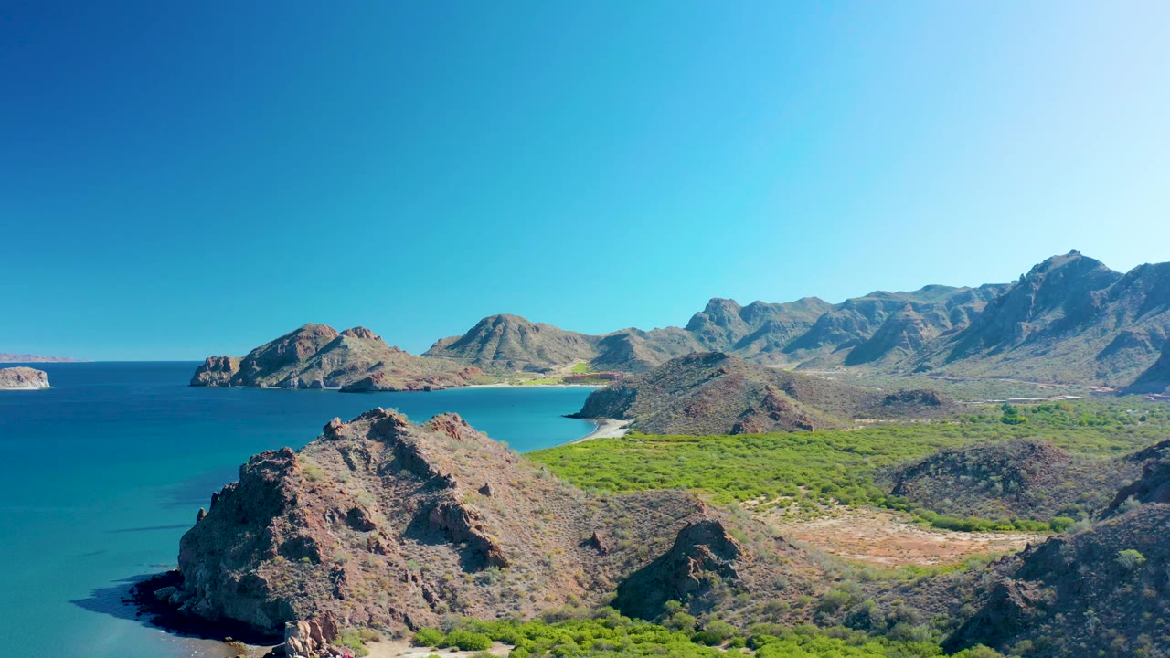 Wide aerial frame of Ligui, Baja California Sur showing bright turquoise water, a rugged rocky point, pale sandy beach, green coastal vegetation, layered mountain ridges, clear sky, and no visible people.