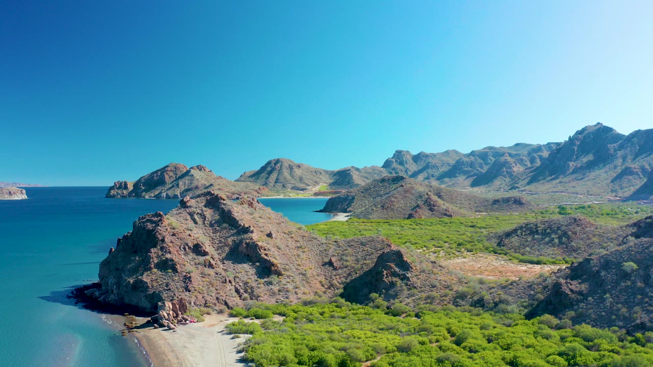 Aerial view of rocky coastline, sandy beach, green vegetation, blue ocean, and mountains.