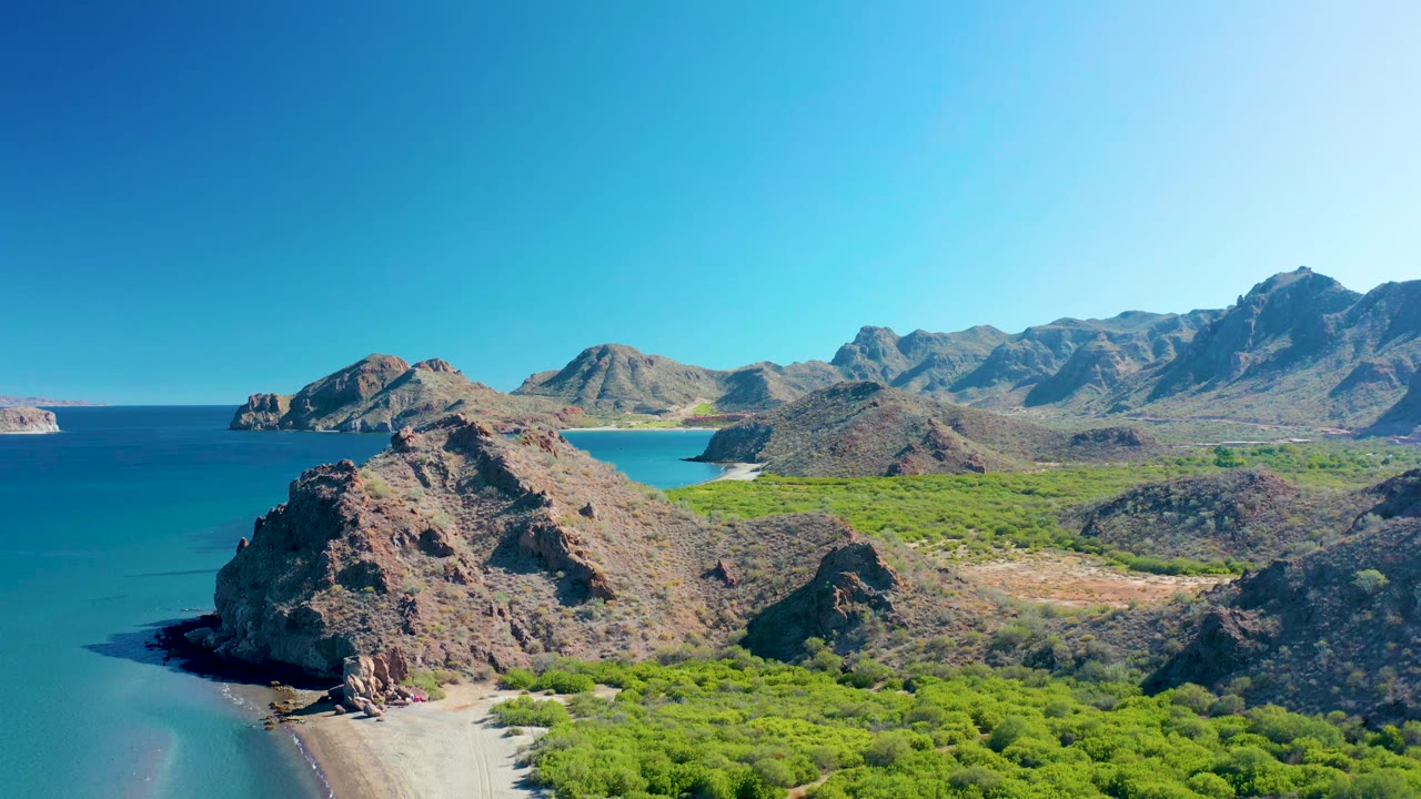Aerial frame in Ligui, Baja California Sur, showing the rocky headland more centered above the curving beach, with blue water to the left, green vegetation to the right, mountains beyond, and no visible people.