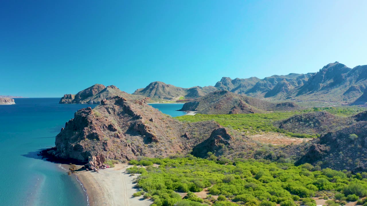Aerial view in Ligui, Baja California Sur, with the rocky point shifted left of center, the sandy beach curving along the lower edge, green scrub filling the right side, mountains in back, and no visible people.