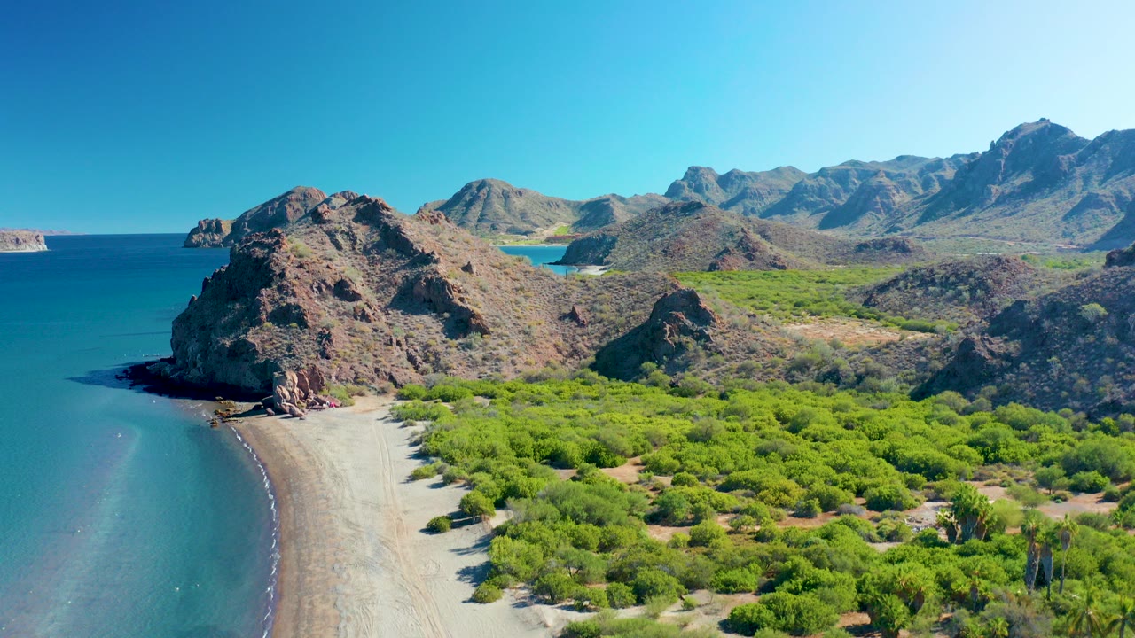 Aerial frame over Ligui, Baja California Sur showing a sandy beach beside blue ocean, rocky cliffs behind the shore, green vegetation in front, and no visible people.