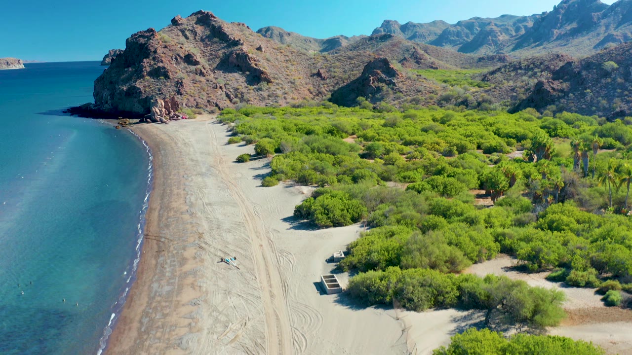 The Ligui shoreline appears from above with calm water at left, a wide sandy strip in the center, bright green trees to the right, and rugged cliffs rising behind the beach.