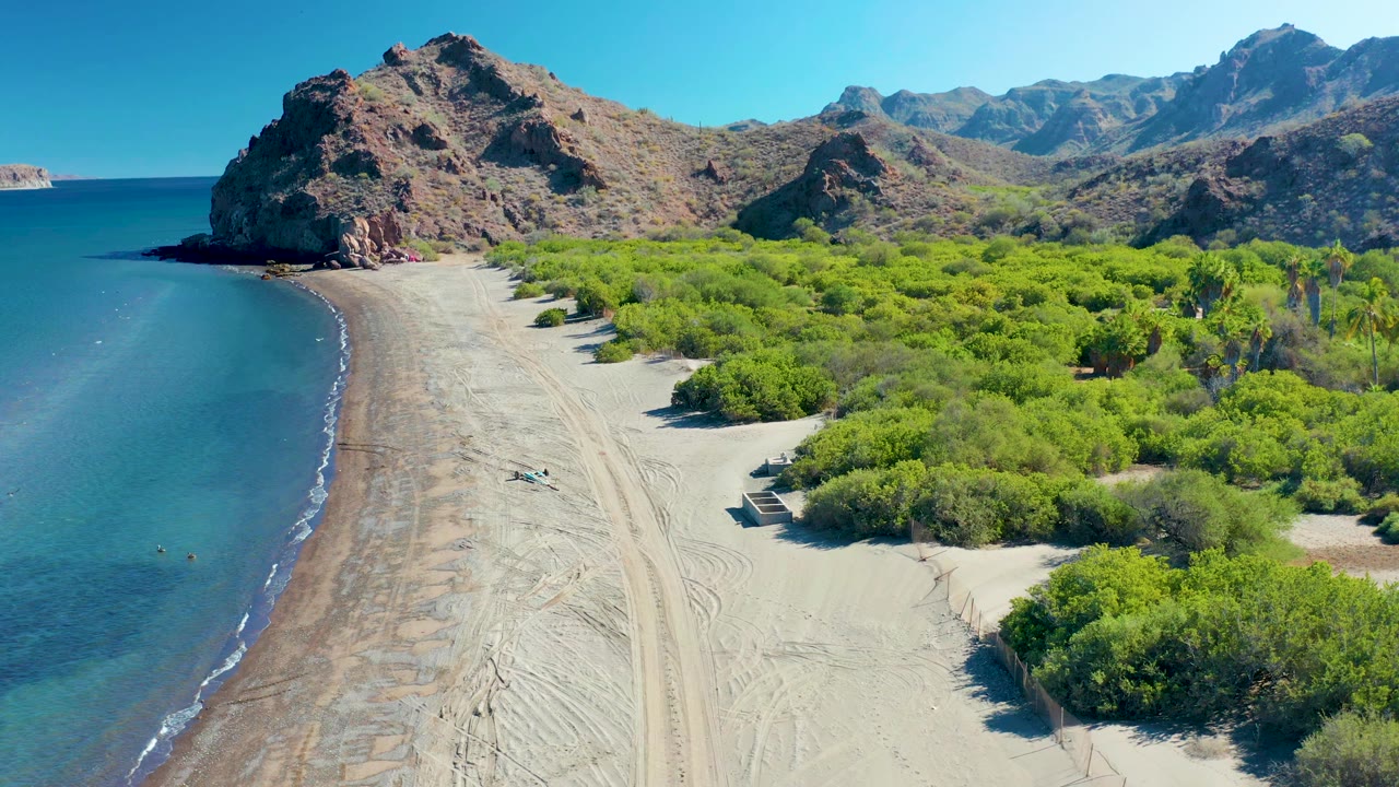 Drone view over a curved beach with ocean, shrubs, mountains, and a small vehicle on the sand.