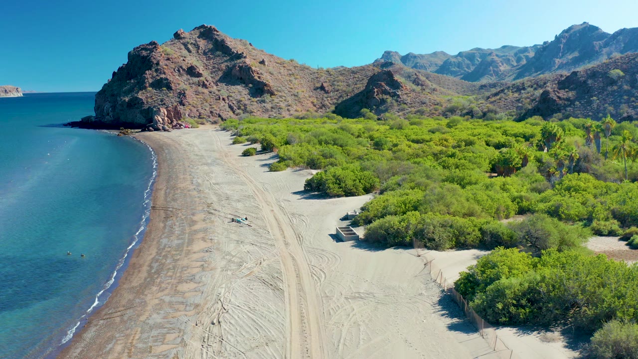 High drone frame over Ligui with calm blue water on the left, a narrow wet shoreline band, a sandy vehicle path through the beach, dense green brush to the right, mountain ridges behind, and no visible people.