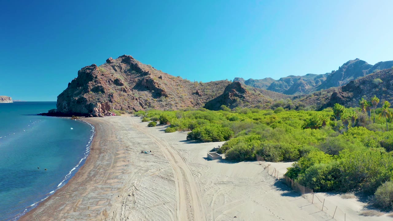 At 0:01, the Ligui shoreline fills the frame with pale sand, blue water along the left edge, bright green shrubs, rocky hills, and no visible people.
