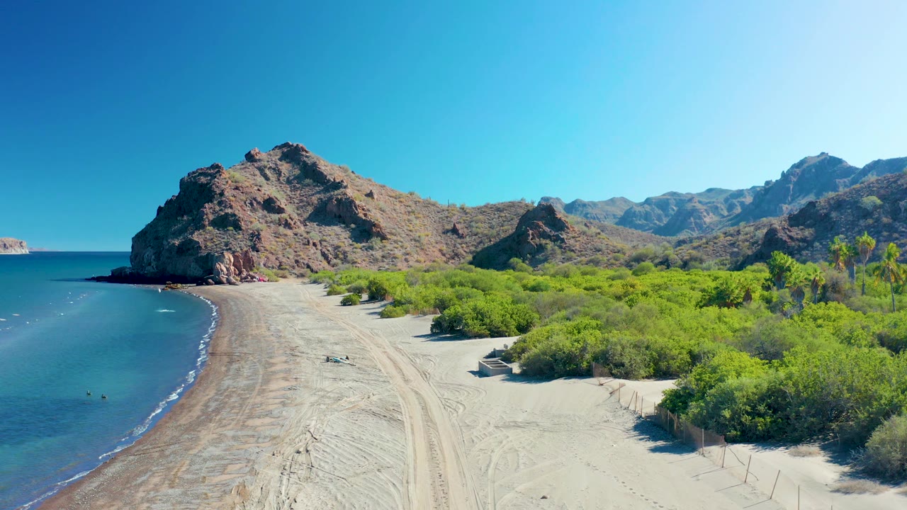 At 0:02, calm ocean borders the left side of the Ligui beach while a rocky headland, low hills, green vegetation, and tire-marked sand remain visible with no visible people.