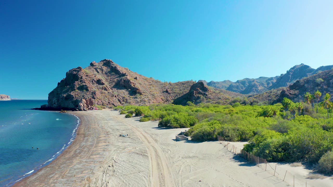 At 0:03, the elevated view shows the Ligui beach widening in the foreground, calm water to the left, shrubs to the right, rocky hills in back, and no visible people.