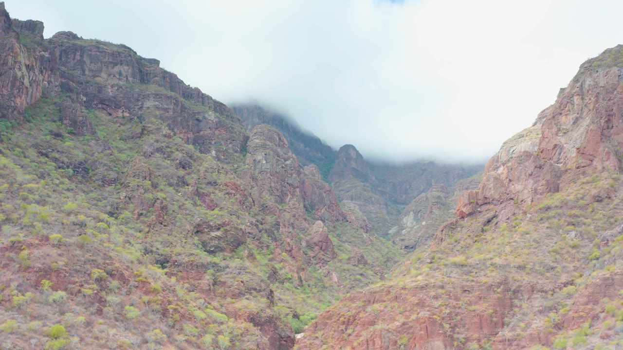 Rocky mountain valley in Puerto Escondido, Baja California Sur, with reddish ridges, sparse green vegetation, low cloud over the peaks, and no visible people.