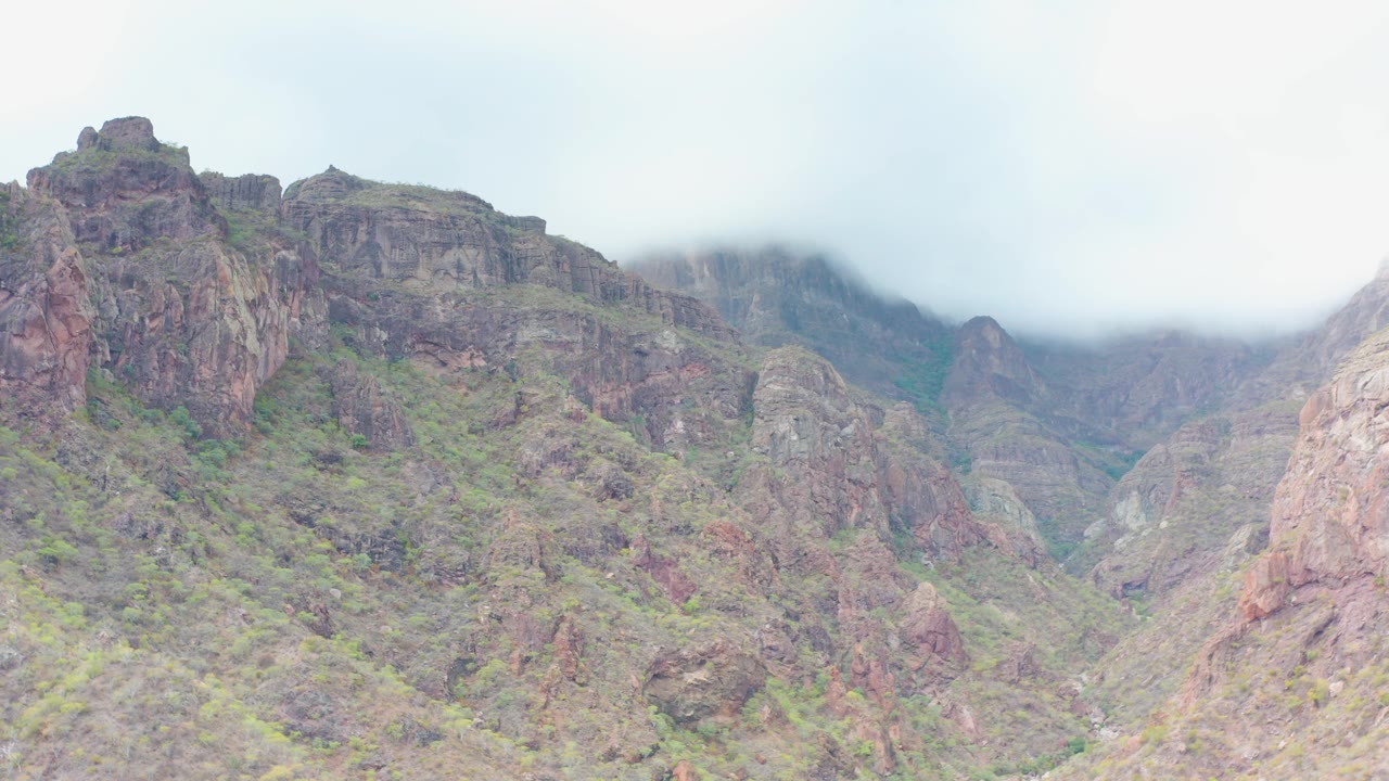 The view holds on jagged Puerto Escondido mountain terrain, where green patches break up the brown rock and low mist covers the upper left ridge, with no visible people.