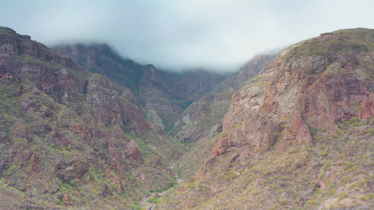 Puerto Escondido, Baja California Sur mountain valley with red-brown cliffs, sparse green vegetation, pale overcast sky, and no visible people at the opening moment.