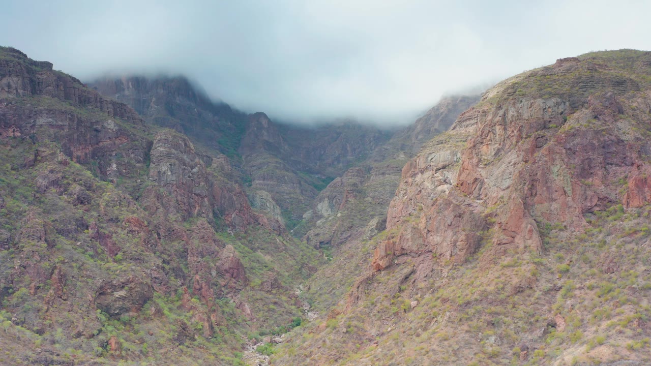 Puerto Escondido, Baja California Sur rocky valley with muted red cliffs, patches of green scrub, a hazy cloud ceiling, and no visible people.