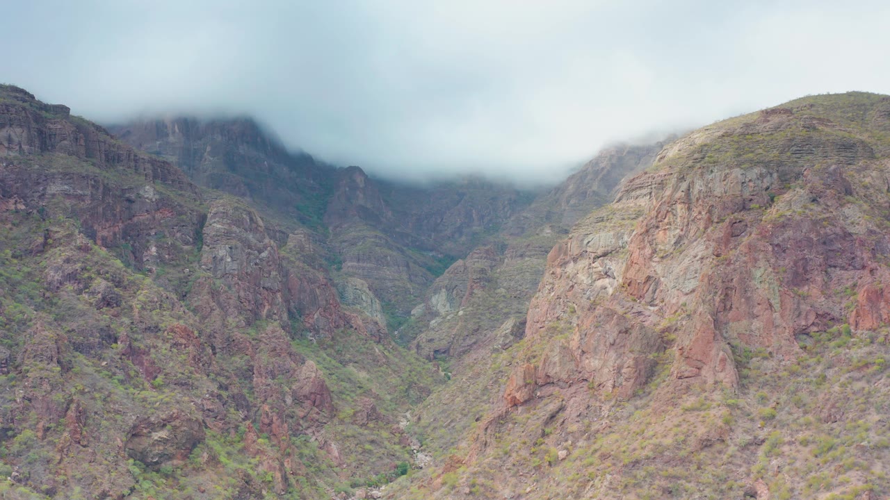 Layered rocky valley terrain in Puerto Escondido, Baja California Sur, Mexico, appears beneath a low cloud bank, with green vegetation in narrow gullies and no visible people.