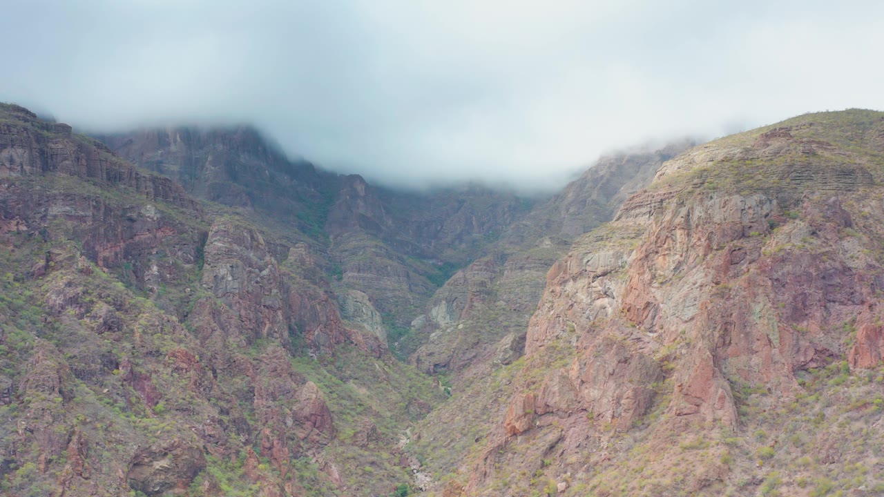 Rugged ridges in Puerto Escondido, Baja California Sur, Mexico, appear under a pale overcast sky, with the upper mountains partly hidden by cloud and no visible people.