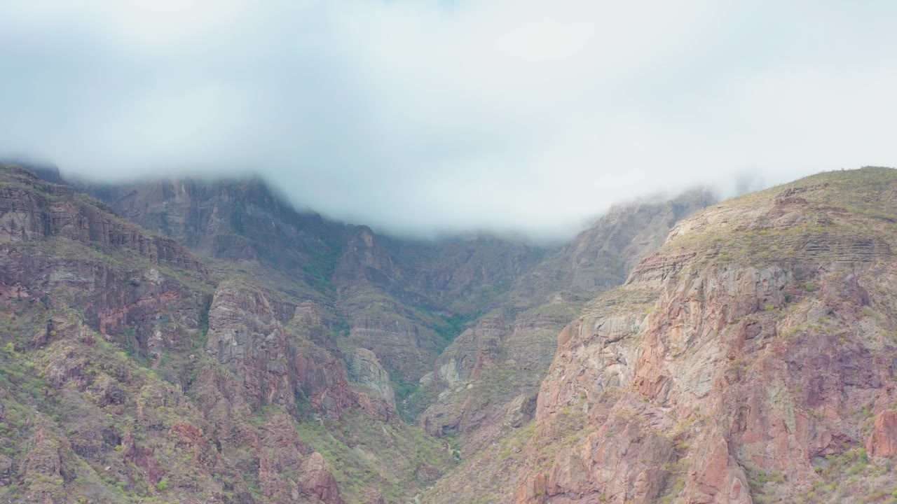 A rocky mountain valley in Puerto Escondido, Baja California Sur shows layered cliffs beneath a thick gray cloud band, with scattered greenery and no visible people.
