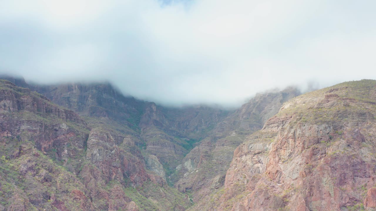 At 0:01, the low cloud layer covers the upper ridges in Puerto Escondido, Baja California Sur, while red-gray rock faces and sparse green growth remain visible below, with no visible people.