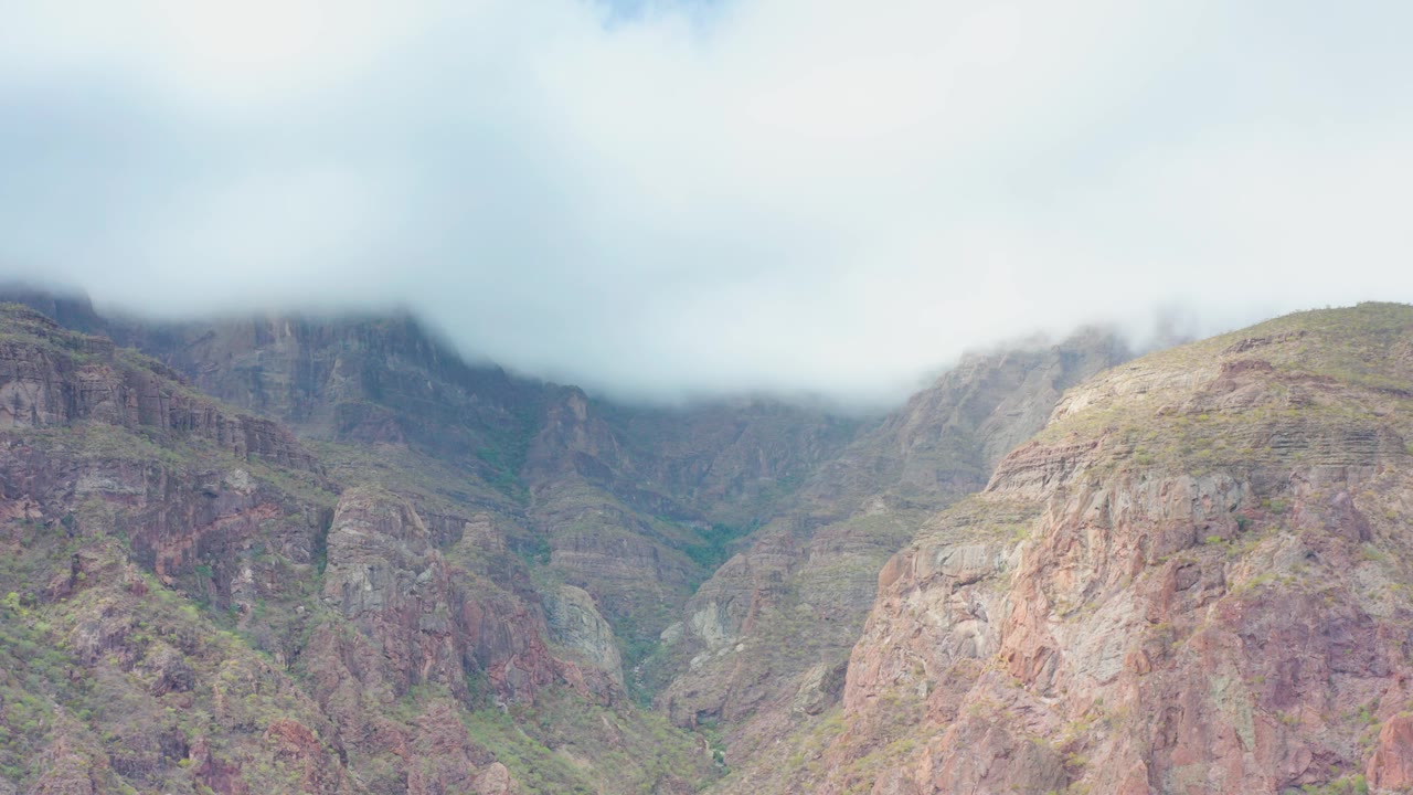 At 0:02, cloud cover partly veils the upper mountain slopes in Puerto Escondido, Baja California Sur, while the central valley and rocky foreground remain visible, with no visible people.