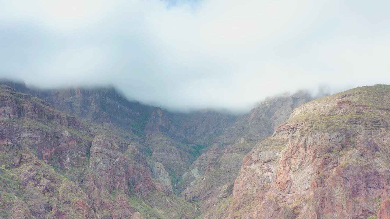 At 0:03, an overcast sky and low clouds sit above a rocky Puerto Escondido mountain valley, with textured cliffs and scattered vegetation visible below, and no visible people.