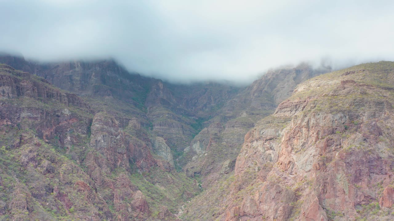 Mist and low cloud press down over the mountain ridge while green gullies cut through rocky slopes in Puerto Escondido, Baja California Sur, with no visible people.