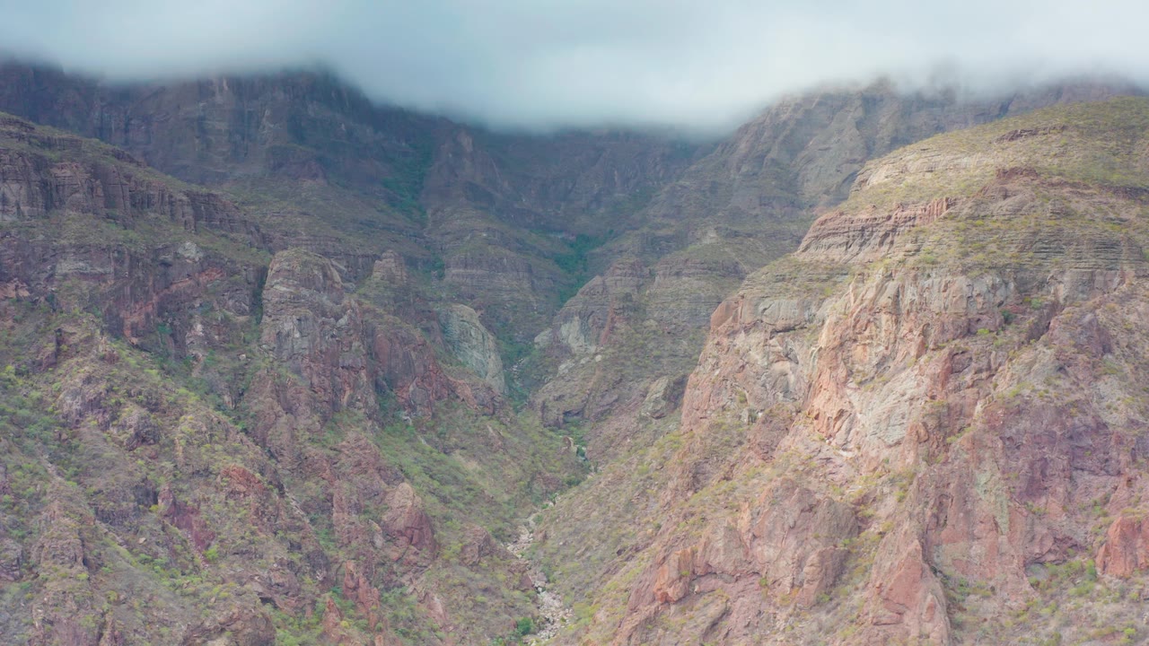 An overcast view shows reddish and gray rock faces, sparse green vegetation, and a narrow valley floor in Puerto Escondido, Baja California Sur, Mexico, with no visible people.
