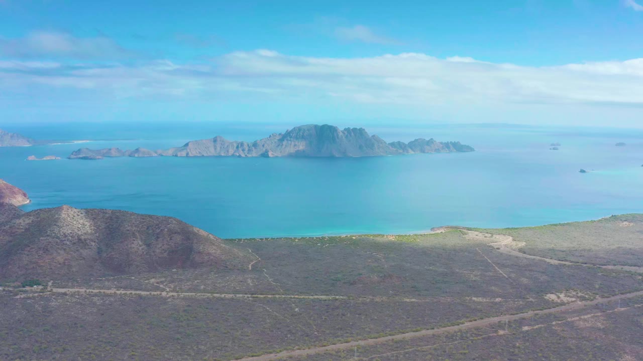 Puerto Escondido aerial frame showing arid scrubland below, a broad blue bay, a rocky island ridge across the horizon, thin cloud cover, and no visible people.