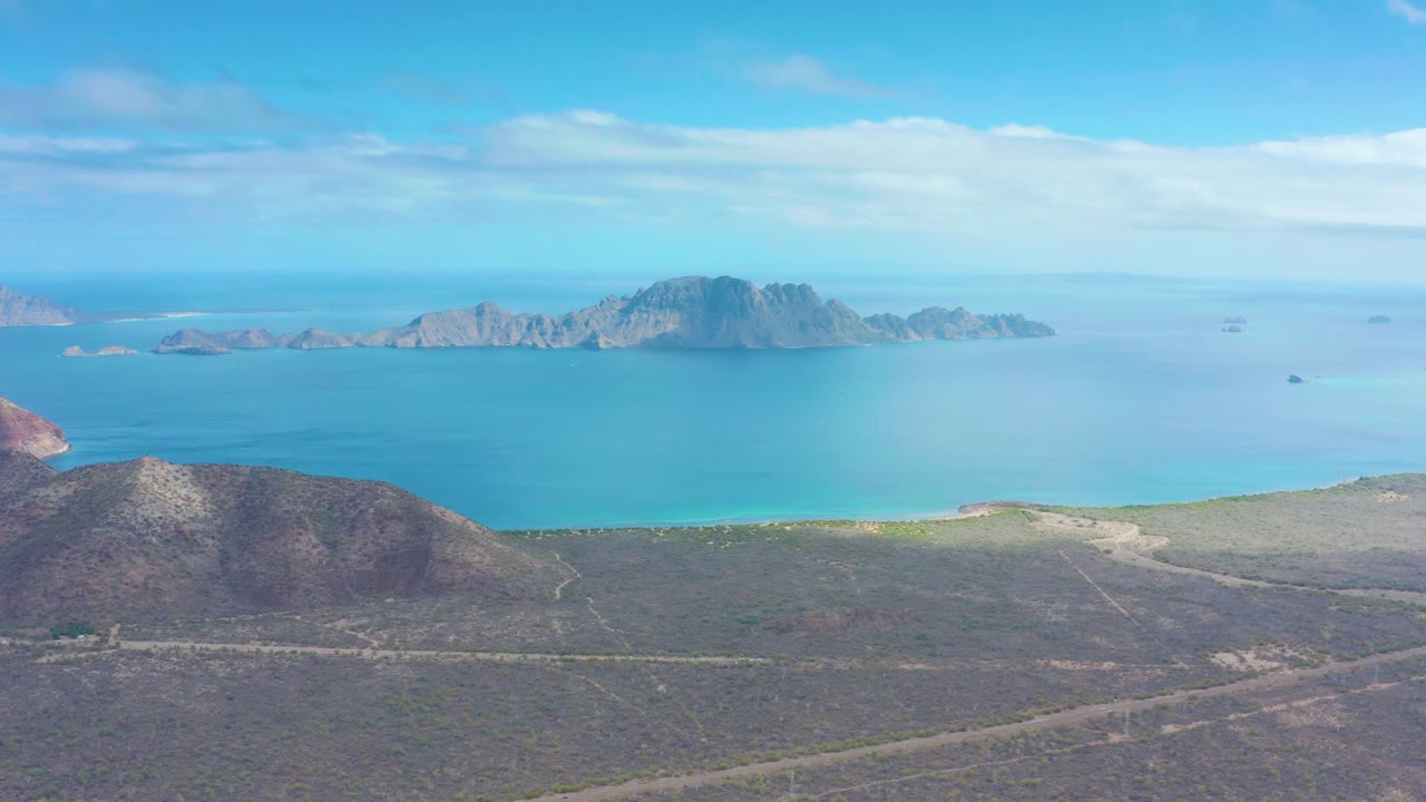 Puerto Escondido coastline seen from above with dry hills in the lower frame, a pale shoreline, calm blue water, offshore mountains, light clouds, and no visible people.