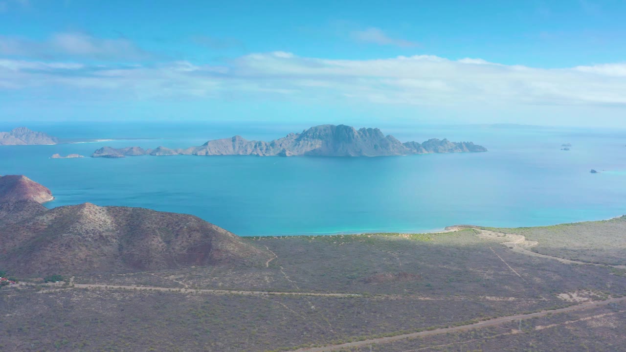 Aerial view in Puerto Escondido, Baja California Sur, showing the dirt road crossing dry coastal terrain while the blue bay and island chain sit across the horizon, with no visible people.