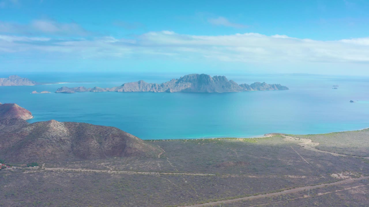 Puerto Escondido aerial frame with rocky foreground hills, sparse vegetation, a pale road near the bottom, bright blue coastal water, distant islands, and no visible people.