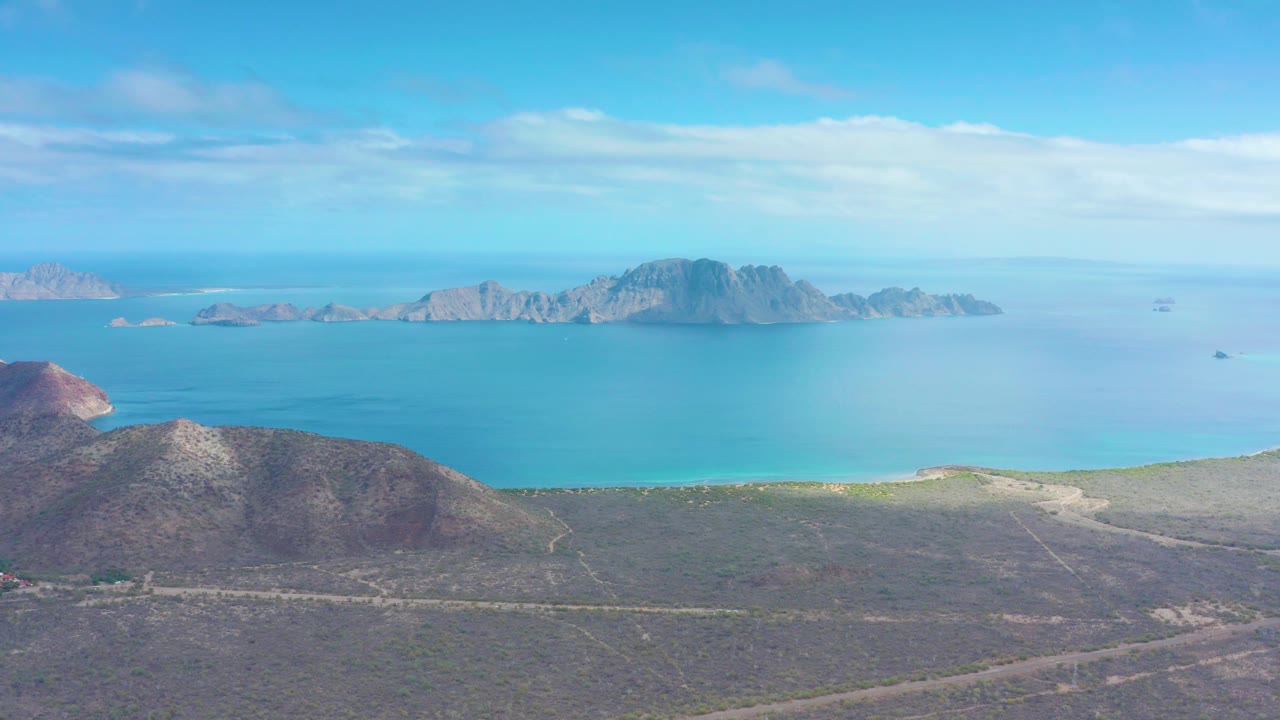Mid-clip aerial frame over Puerto Escondido with a dirt road cutting across dry terrain, a reddish rocky hill at left, blue ocean in back, and no visible people.
