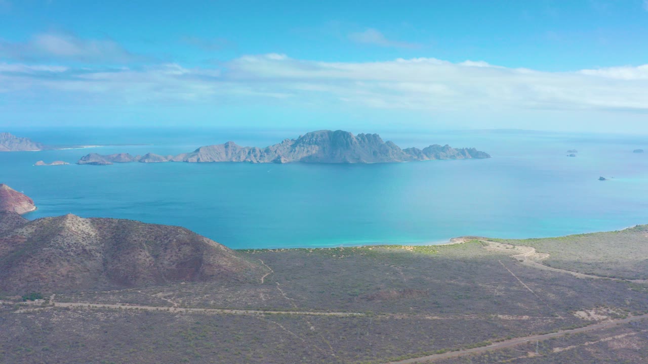 Aerial view in Puerto Escondido, Baja California Sur, showing dry hills in the foreground, turquoise coastal water, distant rocky islands, light clouds, and no visible people.