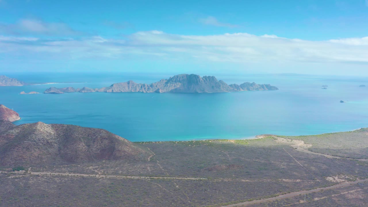 Wide aerial frame over Puerto Escondido with a rounded dry hill at lower left, pale shoreline, blue water, rocky islands on the horizon, and no visible people.