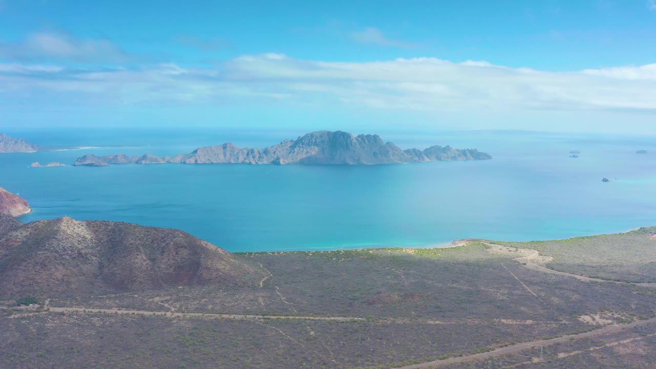 Aerial coastal panorama in Puerto Escondido, Baja California Sur, with arid foreground slopes, clear blue water, rocky island forms, thin cloud cover, and no visible people.