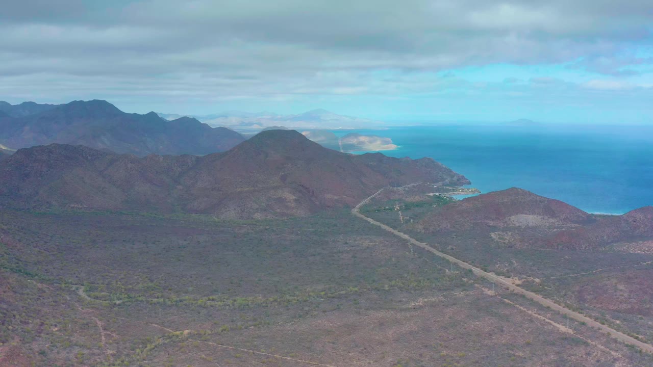 Puerto Escondido aerial frame showing the road angling through brown scrubland toward mountain slopes and a blue coastal inlet, with no visible people.