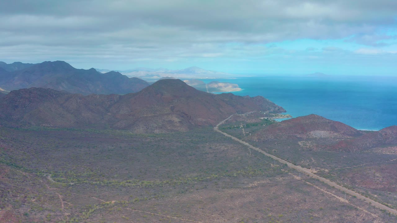 Aerial view in Puerto Escondido with a small vehicle on a light road, scrub-covered desert foreground, layered mountains, blue ocean, and no visible people.