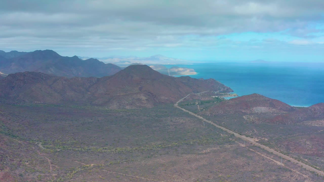 At 0:01, the Puerto Escondido aerial view holds on brown coastal hills, a pale road line, distant blue water, layered mountains, and no visible people.