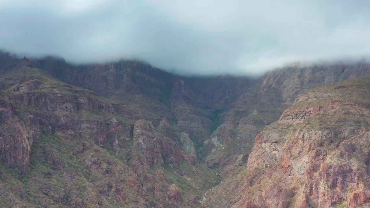 Rocky red and gray slopes fill the Puerto Escondido valley while pale cloud cover presses down across the upper mountains, with no visible people or buildings.
