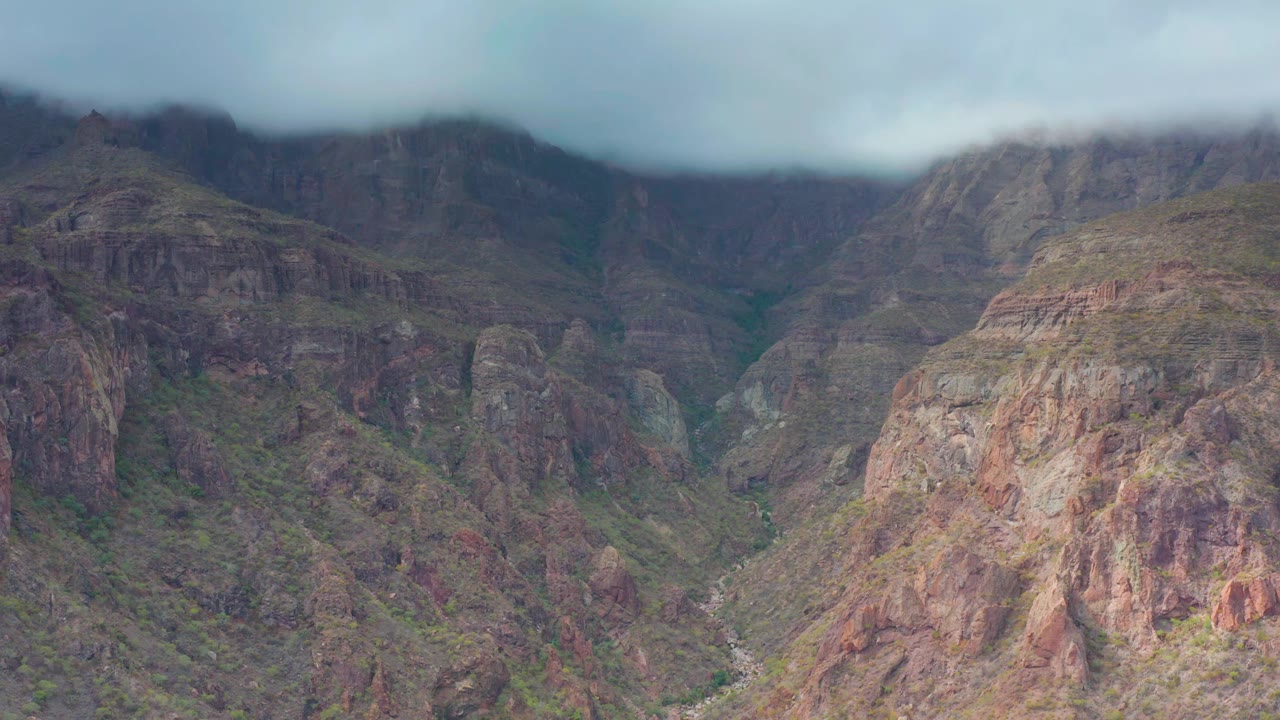 Aerial frame of rugged canyon walls in Puerto Escondido, Baja California Sur, with reddish rock faces, muted green vegetation, a winding pale streambed, and no visible people.