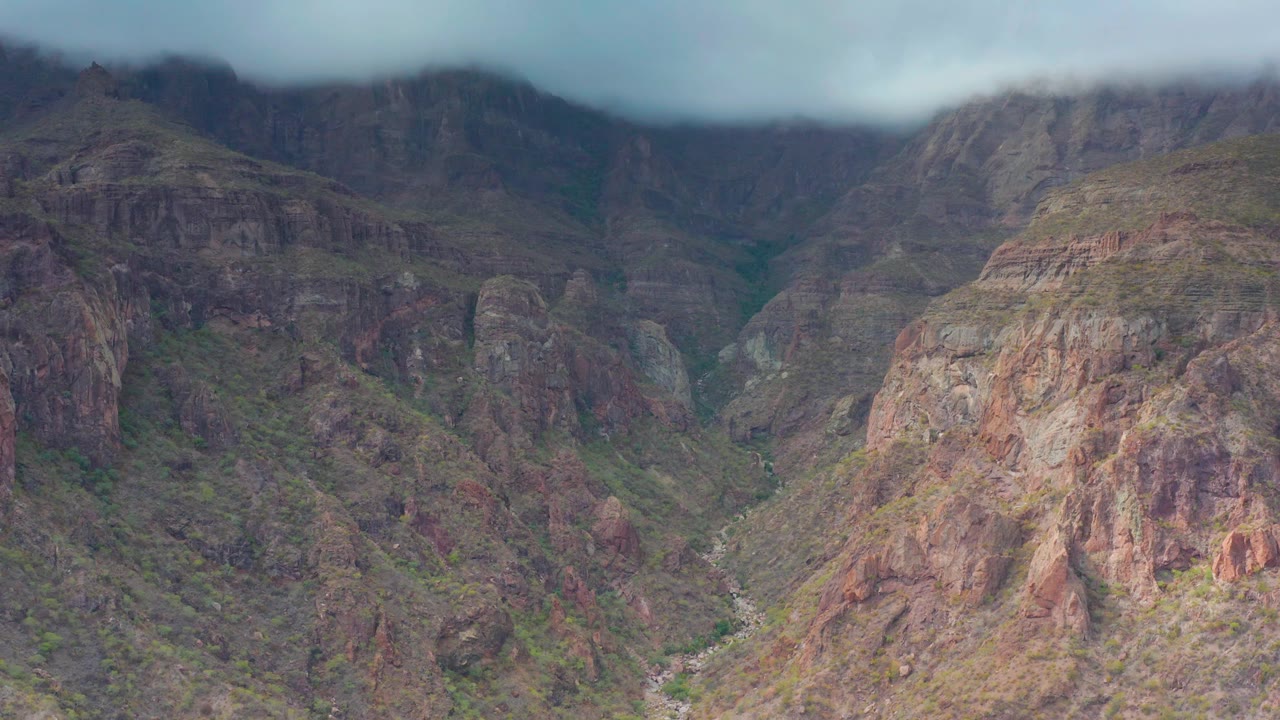 Mid-clip frame in Puerto Escondido, Baja California Sur with a large rounded rock face in front, darker canyon walls behind it, a pale channel below, and no visible people.
