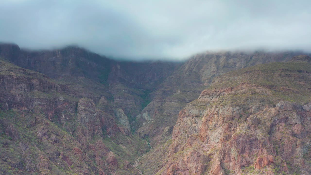 Rugged cliffs in Puerto Escondido, Baja California Sur sit below a soft gray cloud bank, with muted red rock faces, scattered greenery, and no visible people.