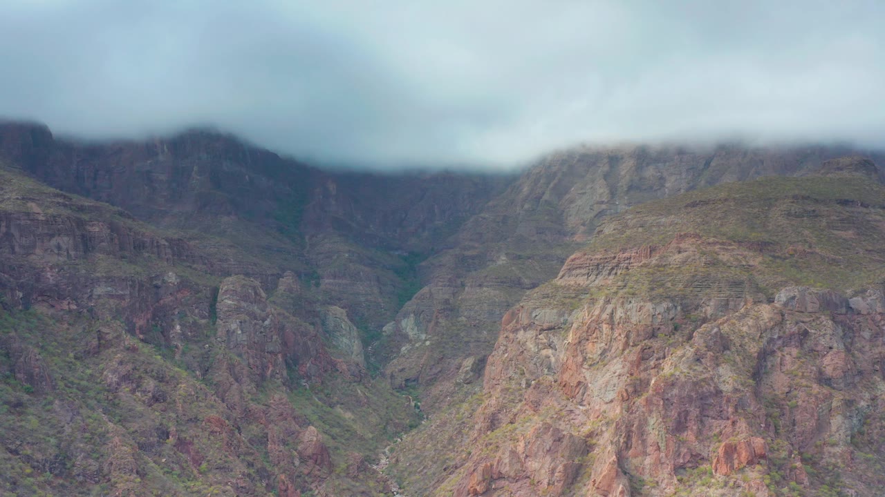 Reddish and gray cliff faces dominate the Puerto Escondido frame, with green streaks in the terrain, a thick cloud layer touching the ridge tops, and no visible people.