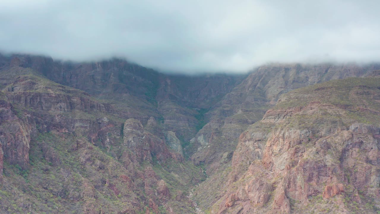A soft cloud bank hangs across the ridge line in Puerto Escondido, Baja California Sur, with reddish rock faces and green valley pockets below, no visible people.